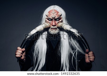 Elderly man holding two red horns. Demon head isolated on black. Ancient alchemy. Devil horns. Bearded old man dressed like Halloween monster. Druid on black background