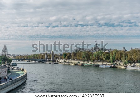 Distinct overview of the sunlight reflections over the Seine river, near the Alexander III bridge and the Grand Palais with many boats, numerous aligned trees and clouds in a blue sky