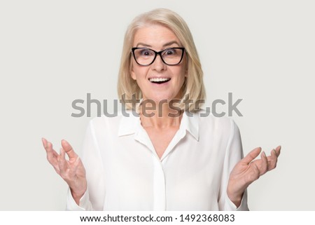 Surprised senior woman in glasses stand isolated on grey studio background feel amazed stunned with good sale offer, shocked mature lady wear spectacles look at camera hold blank copy space place
