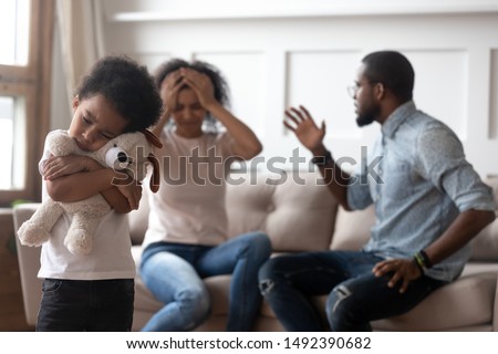 Focus on foreground african little boy feels lonely hugs soft toy suffers from parents scandal, on background sitting on couch mom and dad couple quarrelling scares baby, dysfunctional family concept Royalty-Free Stock Photo #1492390682