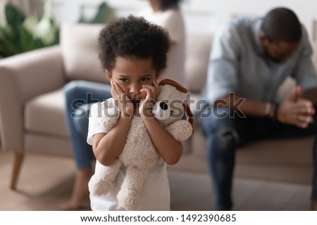 On foreground close up focus on african lonely boy embraces soft toy as symbol seeking for protection support, son closed ears not to hear parents scandal, divorce negative affect to children concept Royalty-Free Stock Photo #1492390685