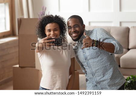Happy mixed race couple capture moment celebrates moving day standing in living room take selfie photo, look at screen wife hold smart phone husband showing thumbs up, big carton boxes on background