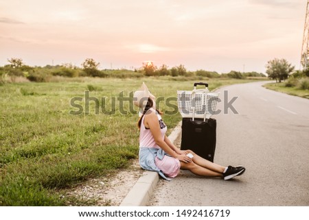 Attractive young girl with a hat on her head and a suitcase by the roadside