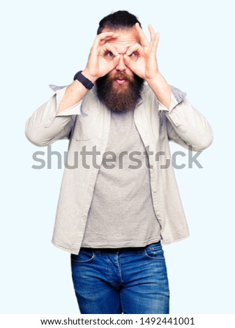 Young blond man wearing glasses doing ok gesture like binoculars sticking tongue out, eyes looking through fingers. Crazy expression.