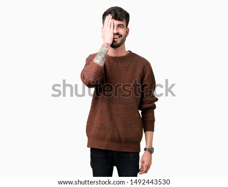 Young handsome man wearing winter sweater over isolated background covering one eye with hand with confident smile on face and surprise emotion.