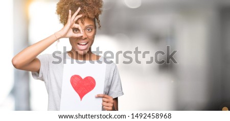 Young african american woman holding paper with red heart over isolated background with happy face smiling doing ok sign with hand on eye looking through fingers