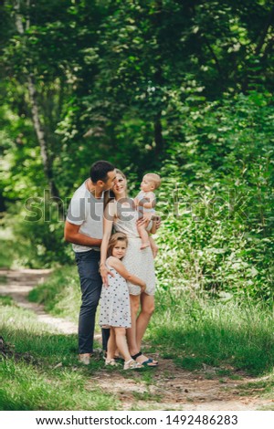 Happy young family spending time together in green nature park. Wife with Husband and two little girls. Mother and father kissing baby daughters.