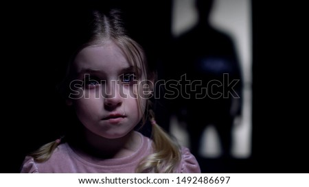 Little crying girl looking at camera, silhouette of rude father outside door