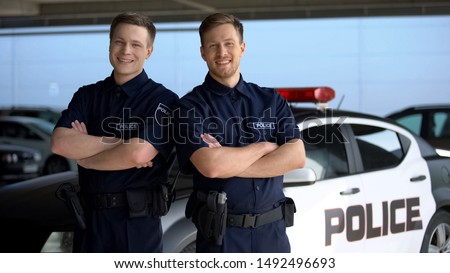Smiling policemen with hands crossed standing against squad car on background