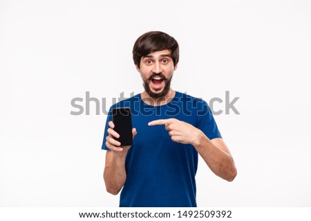 Good looking brunet man in a blue shirt with beard and mostaches shocked looking at the camera pointing at the smartphone standing isolated over white background. Emotion and gesture of surprise.