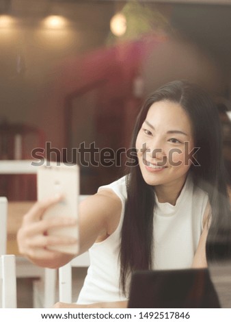 Photo through glass window of beautiful Asian business woman with long black hair sitting in coffee shop with computer on table   smiling and making her own photo with mobile phone.