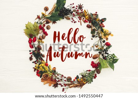 Hello Autumn text, fall greeting sign on autumn wreath of fall leaves, red berries, acorns, anise, nuts, autumn flowers on white wood, flat lay. Seasons greeting card