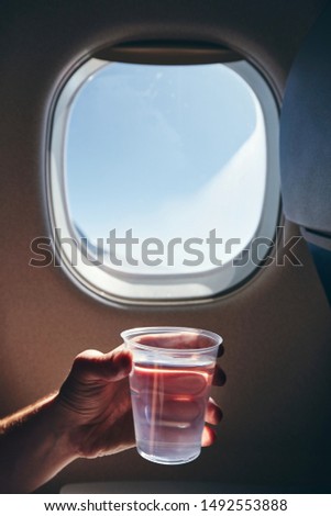 Refreshment during flight. Human hand holding cup of drinking water against airplane window.