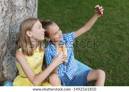 teens boy and girl eat hot dogs sitting on the lawn in the park, have fun, take selfie on a smartphone, lifestyle and summer concept
