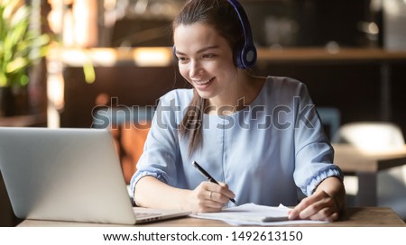Smiling girl student wear wireless headphone study online with skype teacher, happy young woman learn language listen lecture watch webinar write notes look at laptop sit in cafe, distant education Royalty-Free Stock Photo #1492613150