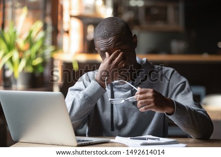 Tired stressed black guy take off glasses feel eye strain after computer work study in cafe, overworked african american business man student suffer from bad blurry vision sight problem sit at desk Royalty-Free Stock Photo #1492614014