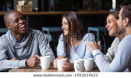 Multiracial friends girls and guys having fun laughing drinking coffee tea in coffeehouse, happy diverse young people talking joking sitting together at cafe table, multicultural friendship concept
