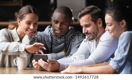 Young diverse friends looking at smartphone using apps in cafe, multiracial people group watching funny mobile social media video on phone together, students mates having fun with cell at meeting
