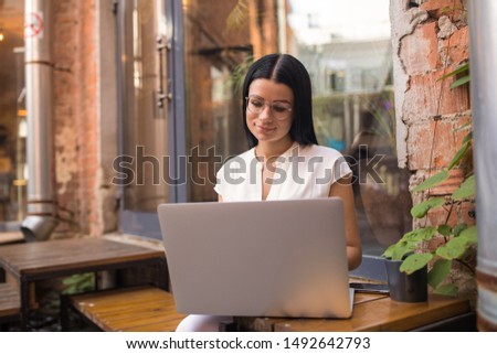 Female in casual wear and fashionable glasses online booking via pc laptop computer while relaxing in restaurant. Hipster girl watching video in social network via notebook, resting in cafe