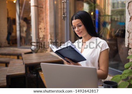Female prosperous business planning expert reading information from diary during distance work on laptop computer while sitting outdoors creative space during recreation time in vacation. 