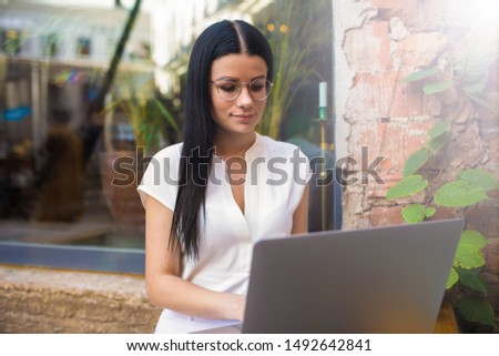 Serious woman in spectacles confident business worker received notifications on laptop computer while sitting outdoors coffee shop. Female graphics designer using netbook, sitting in creative space 
