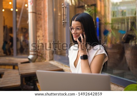 Fashionable dark hair caucasian female in glasses skilled freelance worker talking via mobile phone while sitting outside coffee shop. Woman owner having cellphone conversation after webinar 