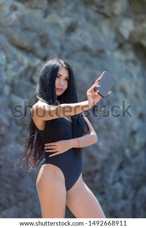 Attractive long haired brunette person resting at secluded place of wild rocky seashore. Dark haired young woman in black one-piece swimsuit takes selfie against coastal rock