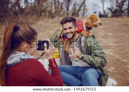 Cute smiling overjoyed mixed race man sitting on blanket and posing with dog while his girlfriend taking picture of them. Picnic at autumn concept.