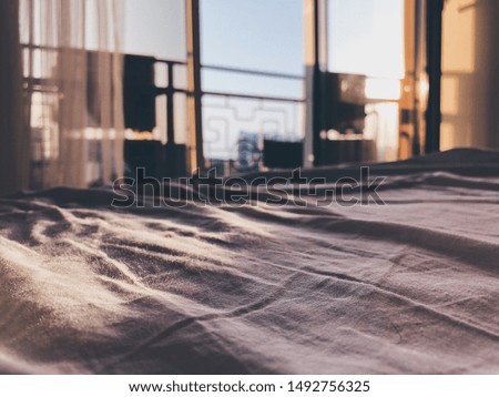 Bed maid-up with clean grey bed sheets in empty room. Close-up. Lens flair in sunlight. Unfocused sunrise city balcony view on the background.