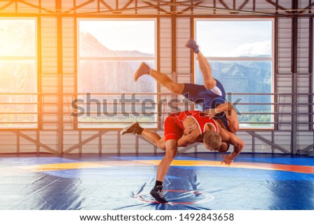 Two strong wrestlers in blue and red wrestling tights are wrestlng and making a  making a hip throw  on a wrestling carpet in the gym. Young man doing grapple.