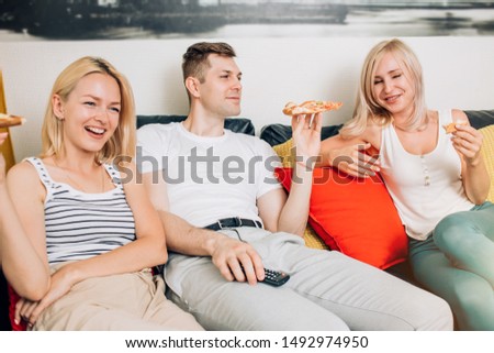 Three young cheerful people eating pizza, laughing, watching funny movie on tv, sitting on sofa in living room at home. Food, leisure, friendship, happiness concept.