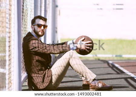 Stylish young bearded guy is sitting on the playground in a suit, tie, snooker and shoes and holding a basketball. The concept of combining business and office work and sports lifestyle.