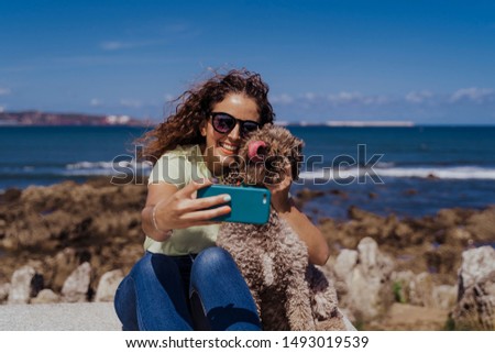 young woman and her cute spanish water dog outdoors enjoying together on a sunny and windy day. Woman taking selfie with mobile phone. Summertime, love for animals and holidays concept
