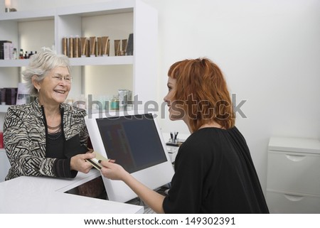Smiling senior woman paying for her haircut through credit card at reception desk