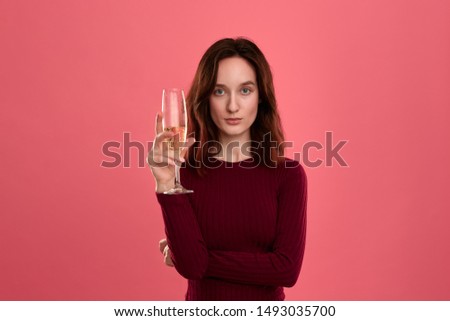 Excited pretty brunette girl holding wineglass of sparkling champagne standing isolated on a dark pink background and smiling at the camera. Celebrating special event.