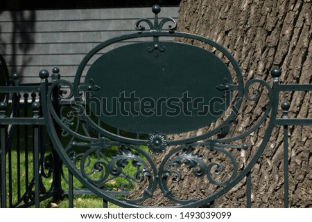 Empty blank sign on metal forging fence in front of big tree. Mock up of information table placard to add any text of symbols. Signboard in park or botanic garden. Element to customize designs