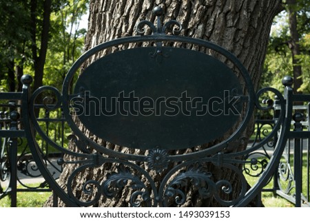 Empty blank sign on metal forging fence in front of big tree. Mock up of information table placard to add any text of symbols. Signboard in park or botanic garden. Element to customize designs
