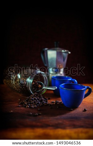 Two Mediterranean blue traditional coffee cups with coffee beans tumbling from a jar. Silver traditional cafetière in the background. Shot on a farmhouse style wooden table against a dark background. 