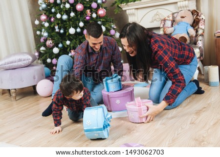 beautiful young happy family caucasian mother father and kid in plaid shirt and jeans sitting near christmas tree and fireplace taking boxes with xmas gifts