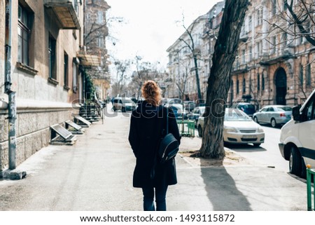 View from behind young woman in black coat and with curly hair walking through an city street next to road and cars on autumn day. Royalty-Free Stock Photo #1493115872