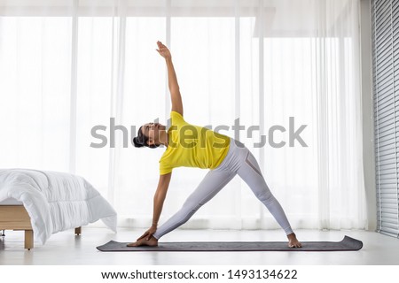 young Asian lady wearing yellow t shirt doing exercise Yoga pose in the bedroom in the morning after woke up. Royalty-Free Stock Photo #1493134622