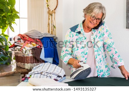 Smiling elderly woman ironing a t-shirt with a lot of clothes waiting for their turn. She talks at cell phone without stops to work. Background of white wall. One caucasian people only
