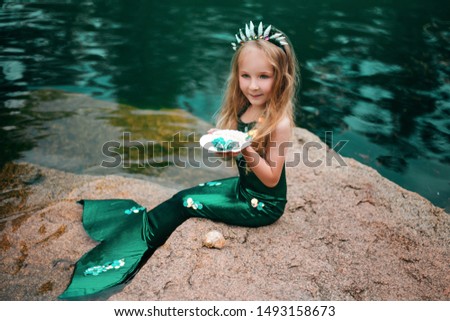 A little girl with white hair with a mermaid tail and shells is sitting on the shore. A little mermaid sits on a large stone by the pond.