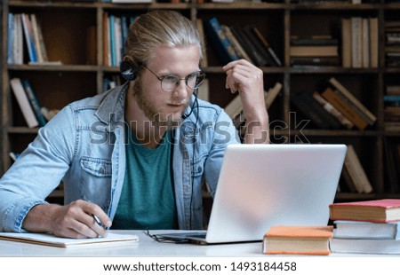 Focused young man student wear headset look at laptop screen make notes study e learning in library watch class webinar training online language course video call distance education software concept Royalty-Free Stock Photo #1493184458
