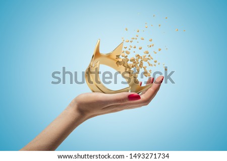 Side closeup of woman's hand facing up and holding gold crown that is dissolving into small pieces on light-blue background. Authority and power. Superiority complex. Visual effects.