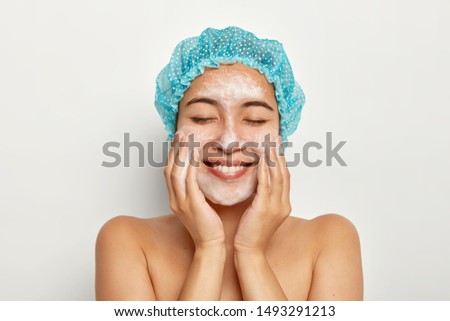 Portrait of delighted young female model applies foaming facial cleanser, touches cheeks, has perfect fresh clean skin after shower, cleans pores, poses against white background. Hygiene concept Royalty-Free Stock Photo #1493291213