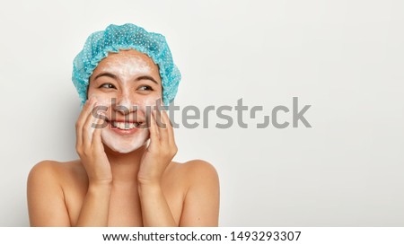 Positive woman applies foam for washing face, wears showercap, smiles positively, stands topless, touches cheeks, looks aside, isolated over white studio wall. Skin care, spa and relax concept Royalty-Free Stock Photo #1493293307