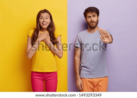 Impressed young woman feels excited and happy, keeps hands on chest, serious unshaven man makes stop gesture, stand closely to each other against colorful studio wall. Emotions, reaction concept