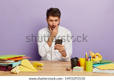 Stupefied young man reads text message, stares with frightened gaze, holds modern mobile phone, works in office, wears elegant white shirt, isolated over purple background. Peope and occupation