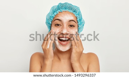 Photo of lovely female model with happy expression, washes face with foaming cleanser, wears wateproof showercap, pampers skin, stands shirtless, looks straightly at camera. Facial treatment Royalty-Free Stock Photo #1493296337